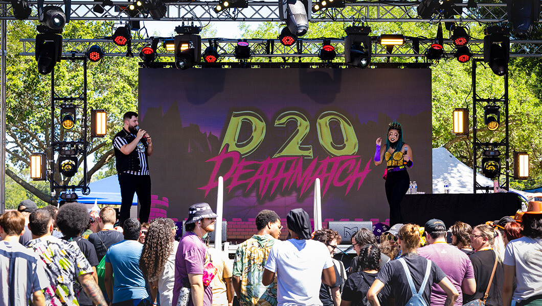 Bec and Drew Godsey wear colorful clothes and stand on an outdoor stage in front of a crowd of students. A screen with the words Deathmatch D20 is behind them.