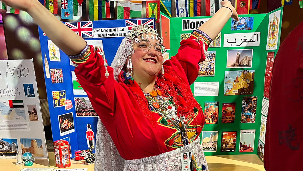 A student wearing a traditional Moroccan skirt, shoes, shirt, and headdress stands with their arms outstretched.