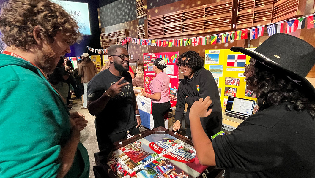 Four smiling students wearing hoodies stand at a table and play a game of dominoes. Displays about different countries are behind them.