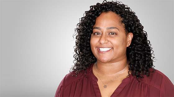 Grad Ambre Moton stands in front of a gray background. She is smiling and wearing a burgundy blouse and a silver necklace with a flower pendant.