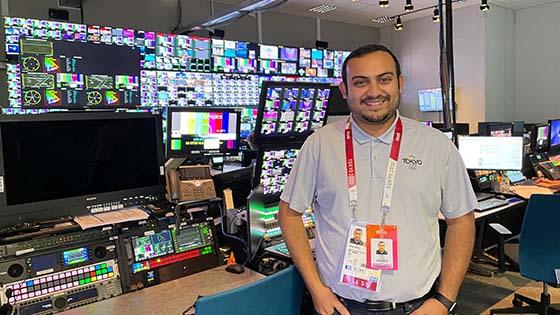 Niraj Patel stands in a broadcast room filled with television screens. He is wearing a gray button-up shirt with a Tokyo 2020 Olympics logo on the chest.