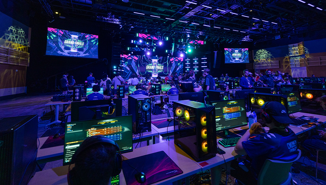 Many competitors sit at consoles in a large room as some bystanders watch the action. Screens display the NACE Grand Finals 2025 graphic.