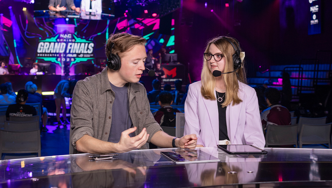 Two sportscasters sit at a table wearing headsets with competitors facing off behind them.