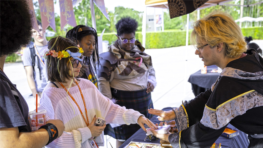 Several students in costume stand in front of a table. One student behind the table holds a deck of cards and another student is picking a card from the deck.