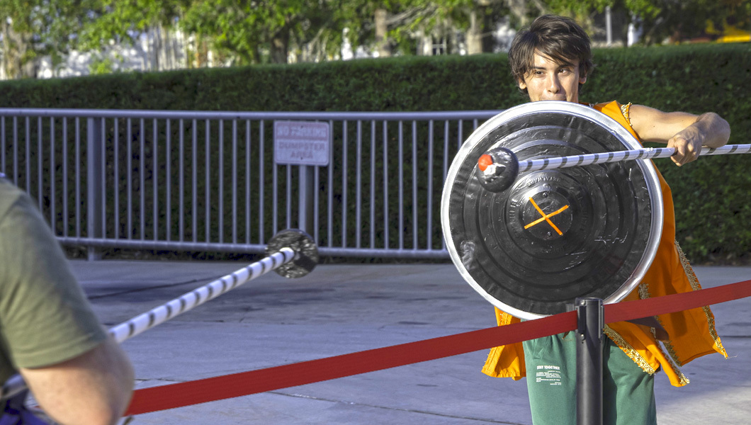 Two students wield lances and shields as they joust. One wears blue armor and the other wears orange armor.