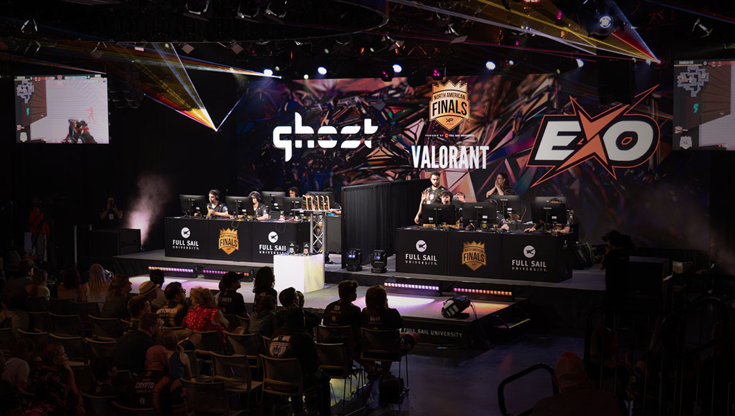 Six players from both Acworth Ghost and Franklin Exo sit at gaming consoles onstage with a black curtain between them as an audience watches. A large screen displays the XP League North American Finals graphic, as well as the game they’re playing, Valorant.