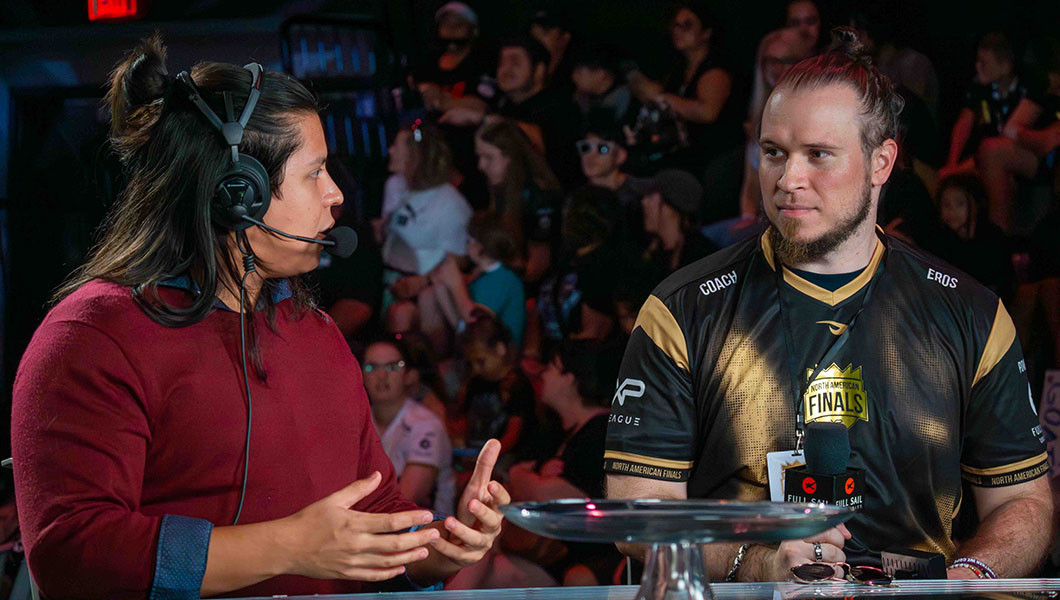 Wearing a headset and a red sweater, PB_n_John speaks to Coach Eros who is wearing a jersey and holding a microphone. A crowd sits on bleachers behind them.