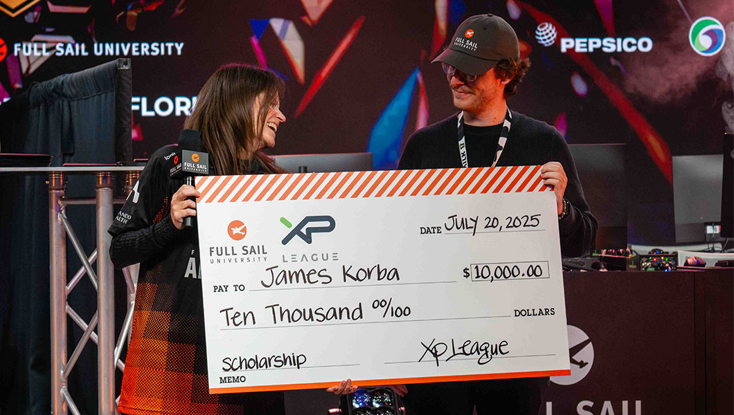 Sari Kitelyn smiles as she hands a giant $10,000 check to James Korba. Sari wears an Armada jersey and holds a microphone, while James wears a Full Sail cap, lanyard, and black shirt.