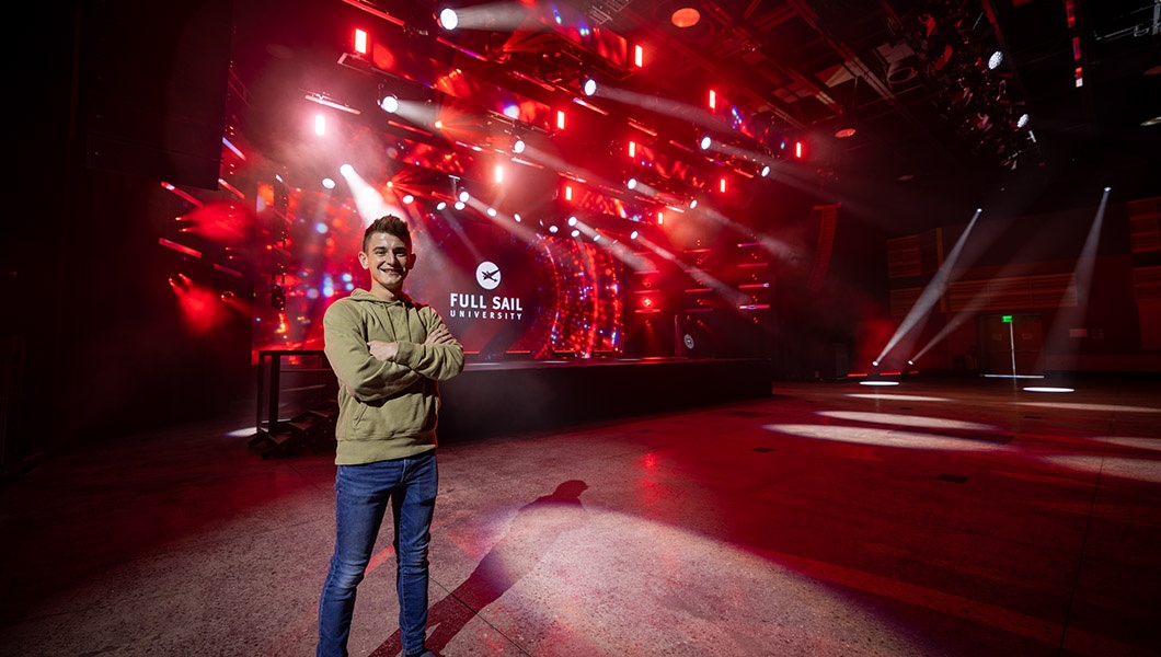 A smiling young man wearing jeans and a hoodie stands with his arms crossed in front of a large LED screen in Full Sail’s live events venue.