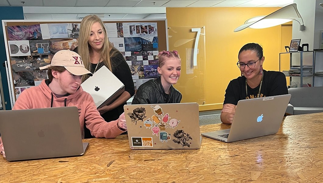 Two students using laptops sit at a table with an Art Director. A second Art Director stands behind them.
