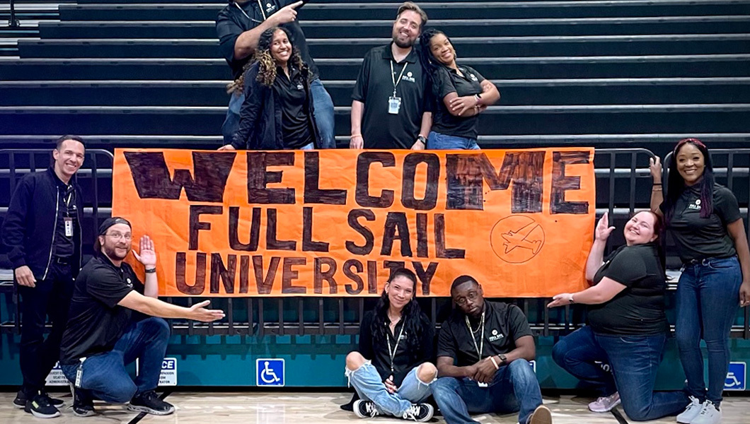 Full Sail staff members posing for a photo with an orange handmade sign that reads “Welcome Full Sail University.”