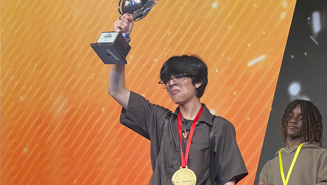 A young man on stage holding up a silver trophy over his head while wearing a large gold medal around his neck.