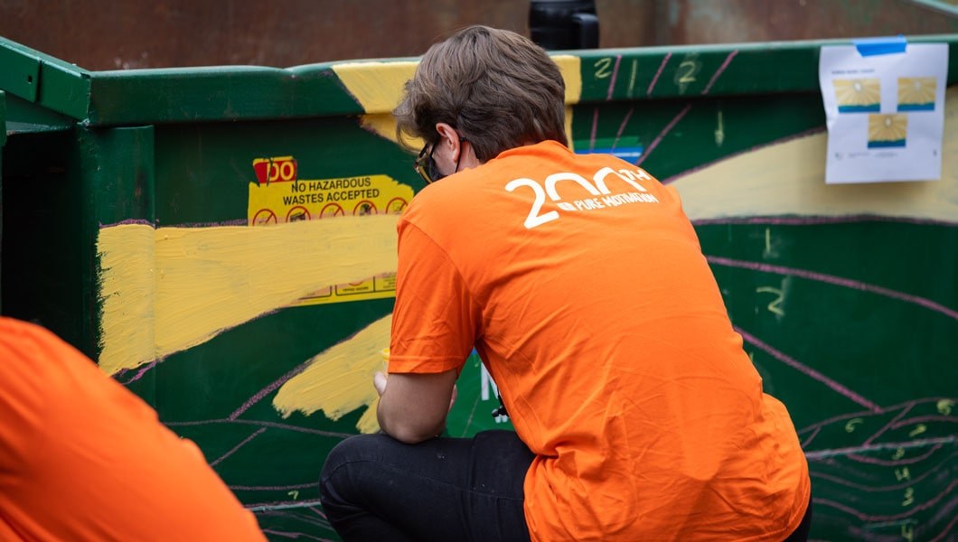 Man wearing a bright orange t-shirt crouches down with his back to the camera as he paints a large green trashcan.