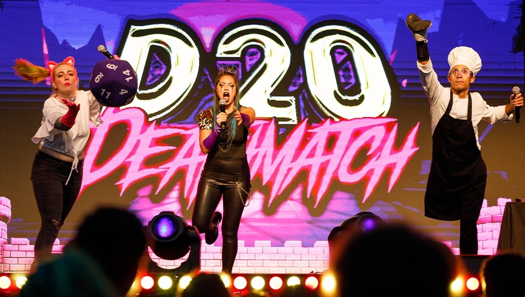 Three people on stage in make-up and costumes rolling a giant 20-sided die, the logo for D20 Deathmatch appears on the screen behind them.