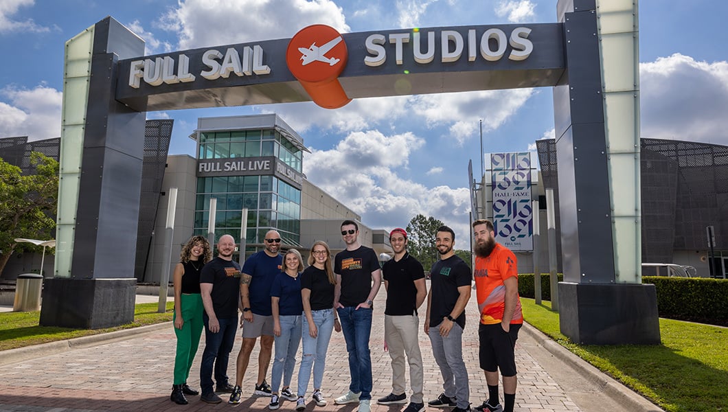 A group of nine individuals smiling and standing underneath an archway that reads ‘Full Sail Studios’ on a bright, sunny day.