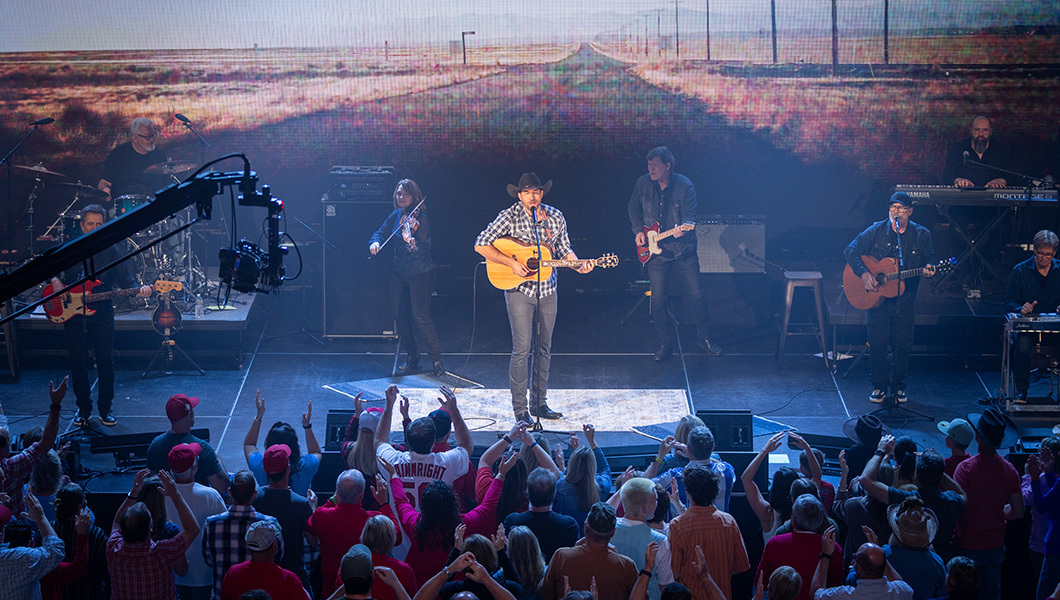 A man wearing a cowboy hat and flannel shirt is seen center stage while playing the guitar with an ensemble band in the background.