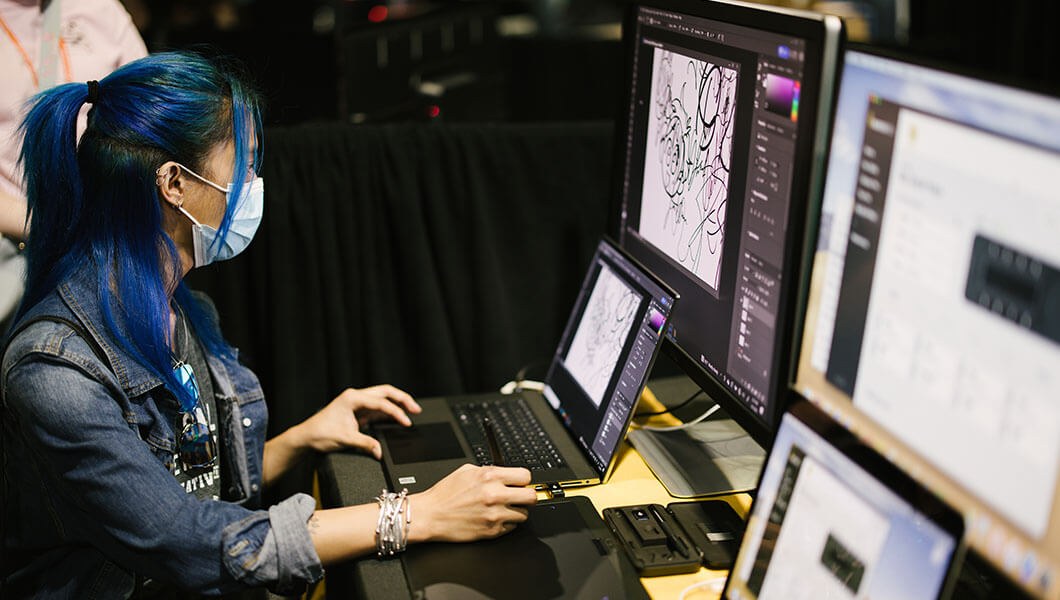 A woman wearing a face mask, and a t-shirt with a jean jacket over it sits in front of a laptop and desktop monitor. The screens are displaying drawings in Adobe Photoshop.