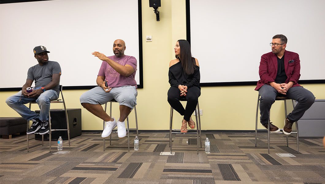 Two men wearing jeans and t-shirts, a woman in black pants and a black blouse, and a man in jeans and a blazer sit in front of two whiteboards in a classroom.