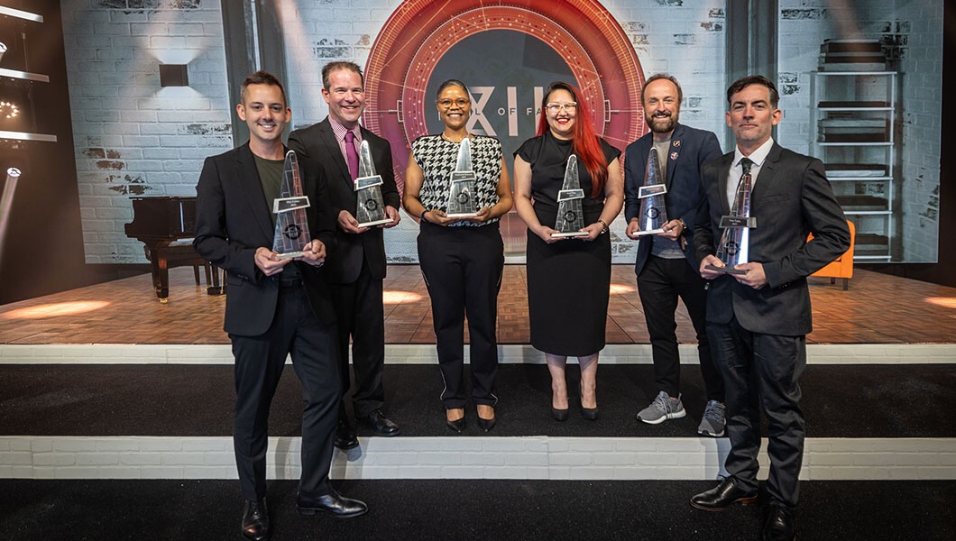 Four men and two women stand in front of a stage with a backdrop saying Hall of Fame XII. They are all holding triangular silver statuettes.