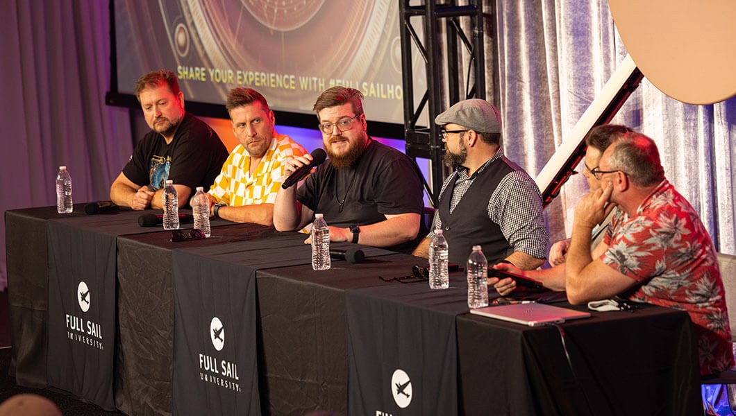 Six men sit at a long table draped in black fabric sheets bearing the Full Sail logo. There is a screen behind them with the words Share Your Experience With #FullSailHOF on it.