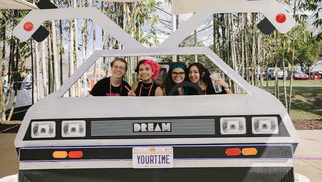 A smiling man and three smiling women sit inside an outdoor photo booth. The booth is shaped like the Delorean from ‘Back to the Future,’ with a license plate that says Hall of Fame XII YOURTIME.