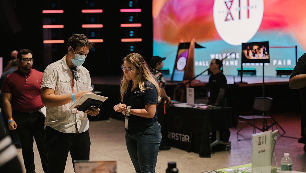 A man holding papers and a tablet speaks with a woman. They are standing in a room with tables representing different employers and a large screen that says Hall of Fame XII in the background.