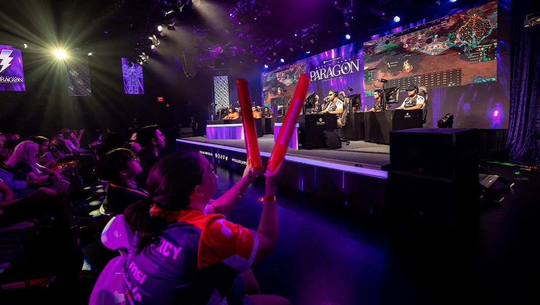 Several competitors sit on-stage at desks in front of large monitors looking very focused. The crowd is cheering.