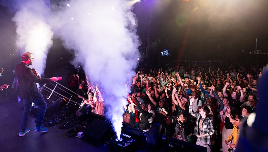 A man in a black blazer is throwing prizes into the crowd. There are two smoke machines on stage. The audience is smiling and they are reaching towards the front of the stage to catch the prize.