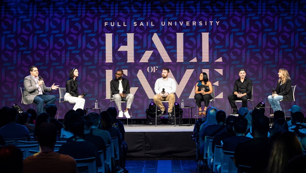 The Hall of Fame inductees are sitting in a line on the stage with a moderator in front of a giant screen that reads: Full Sail University Hall of Fame. They are all holding microphones and look happy to be there.