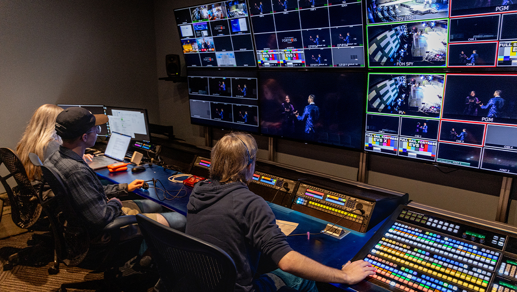 Three students sit at a control panel with multi-colored keys and large screens.