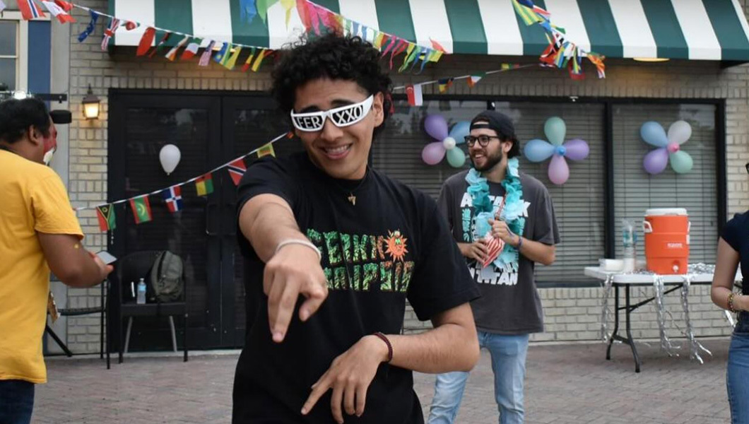 A student wears sunglasses and dances under a banner of flags of various countries. Other students stand around.