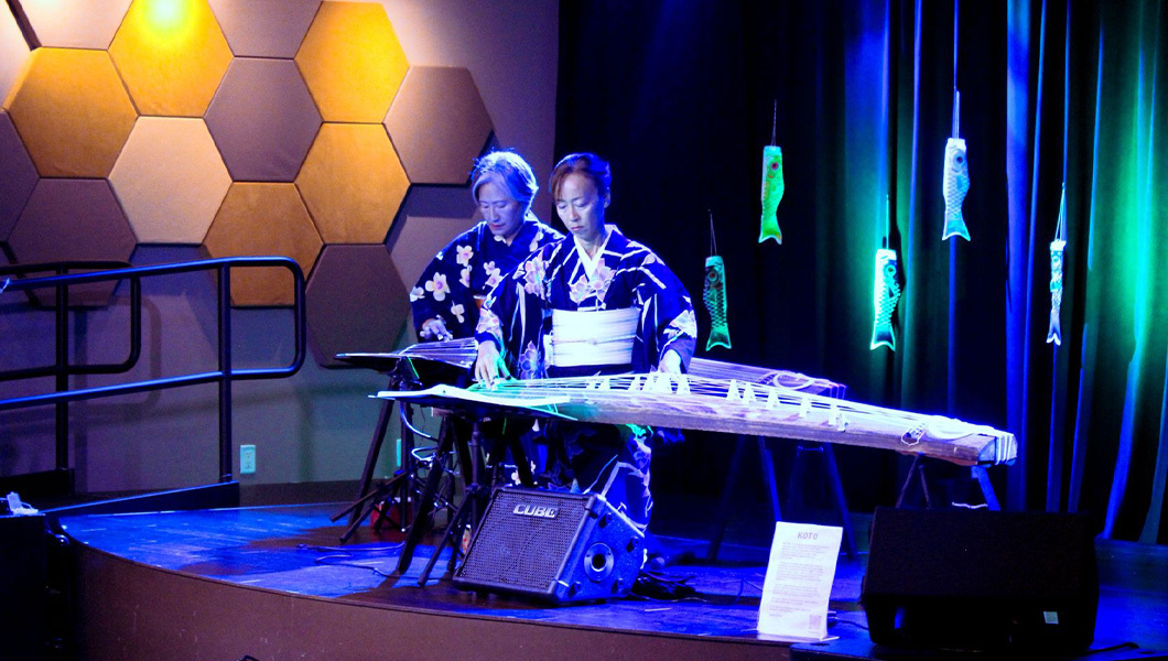 Two people dressed in Japanese kimonos play the koto side by side on a stage. Paper fish lanterns hang behind them with green and blue lights.