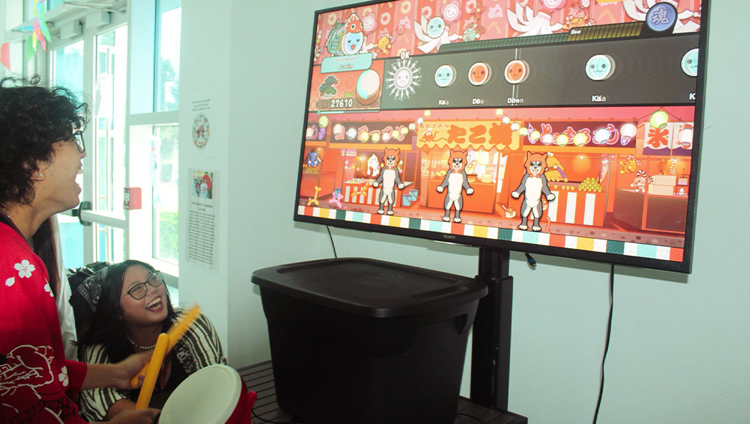 A student plays a toy drum video game facing a screen displaying the game. Another student sits nearby watching.
