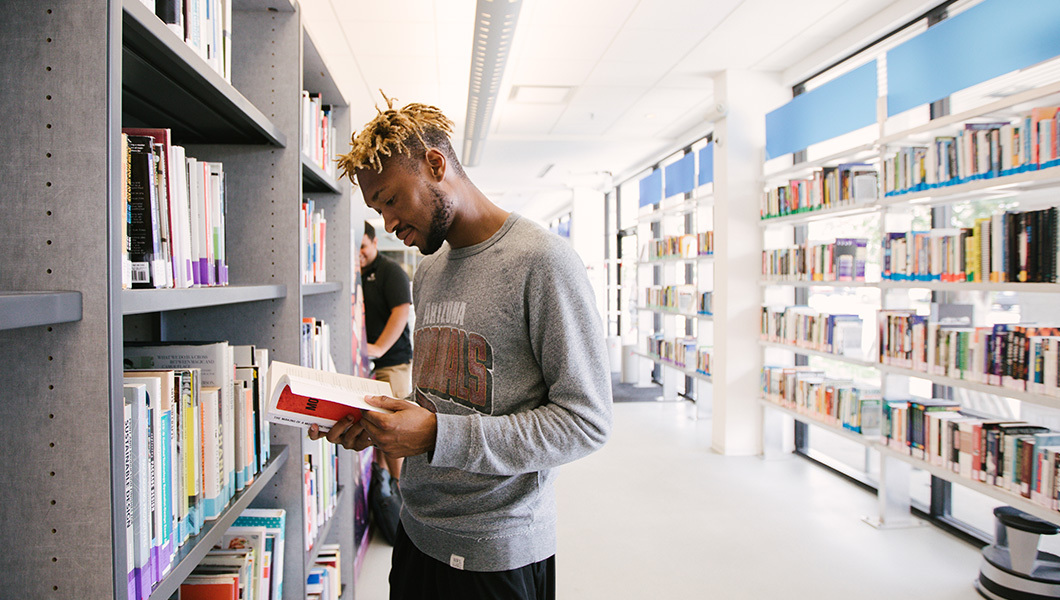 A college student reads a book in a sunny aisle lined with shelves in a quiet, modern library setting.