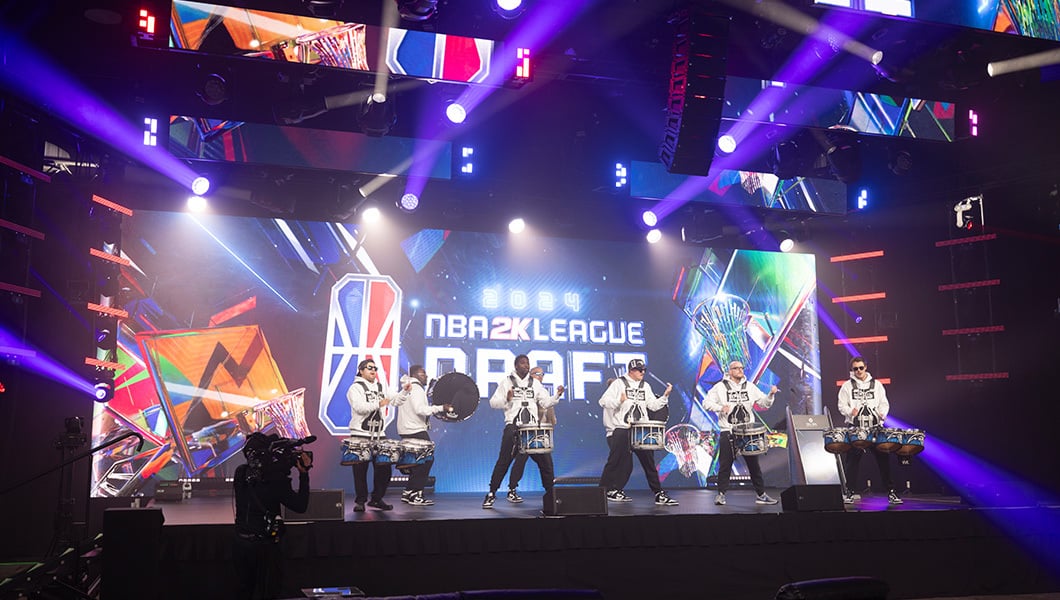 A drumline is performing on a stage in front of a large LED screen that reads “NBA 2K League Draft.”