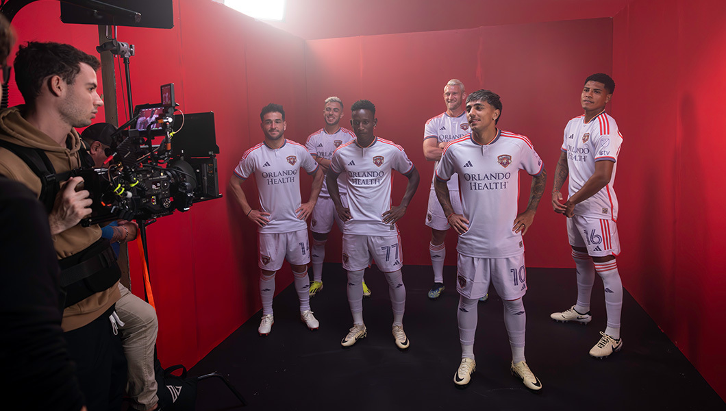A group of six soccer players in full uniforms pose in a production studio while being filmed against a red backdrop.