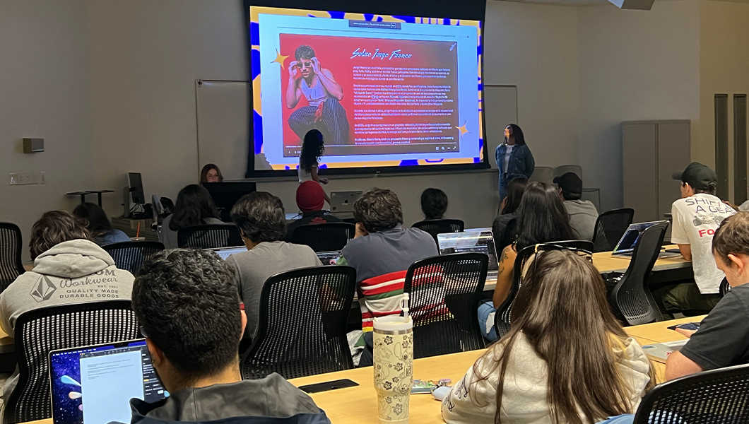 Sofia Adamary and Gabriella Pedraza stand at the front of a room full of students sitting at desks. Several students have their laptops out. A projector screen behind Sofia and Gabriella displays “Our Vision & Structure.”