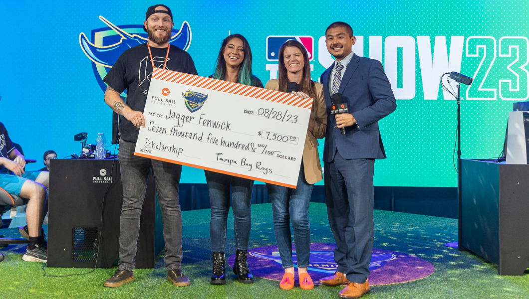 A group of four people standing and smiling on a grass-covered stage while holding a large scholarship check.