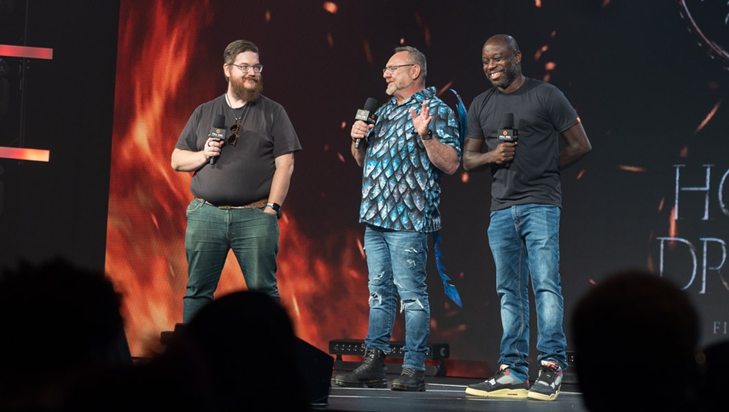 On stage inside the Full Sail Live Venue, Stephen Beres, Rick Ramsey, and Leslie Brathwaite hold mics and stand before a large screen showing the fiery House of the Dragon logo.