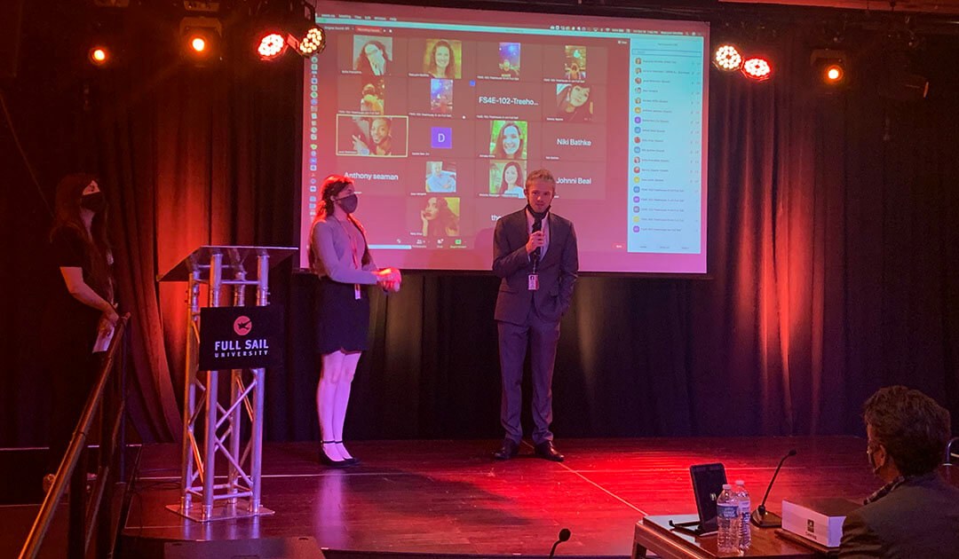 A woman wearing a skirt and sweater and a man wearing a suit stand on a stage in front of a competition judge while another woman wearing black looks on. There is a lectern with a Full Sail University sign on the right side of the stage.