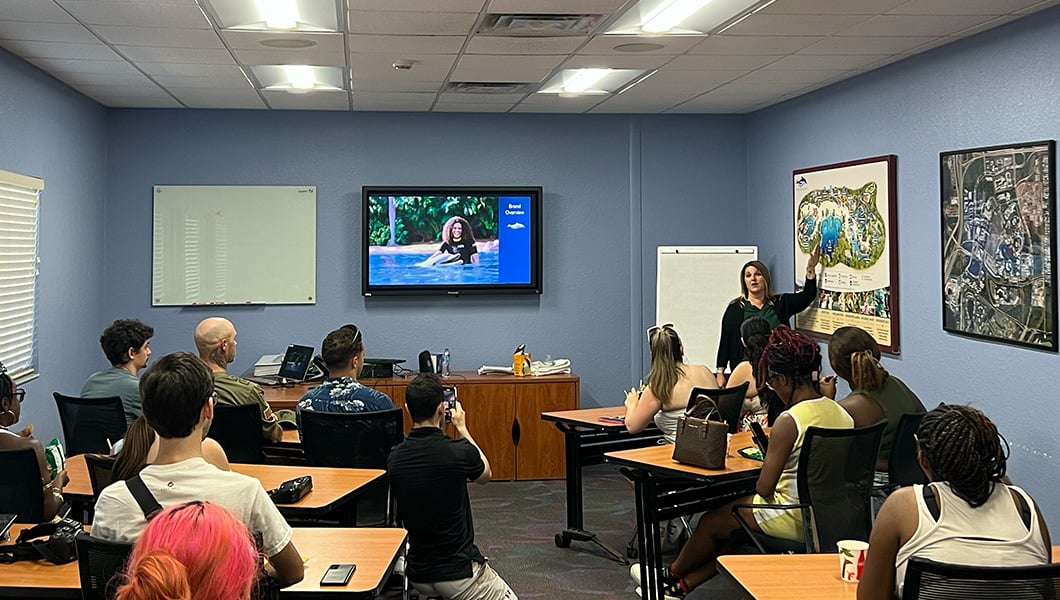 Students sit at desks as Crystal O’Hea stands at the front of the room and speaks. There is a screen on the wall displaying a slide presentation.