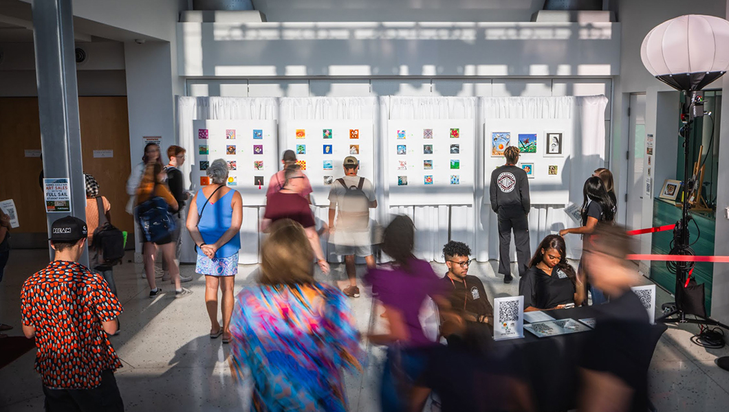 A long exposure image of people staring at the many small paintings hung up on the Full Sail Live lobby wall.