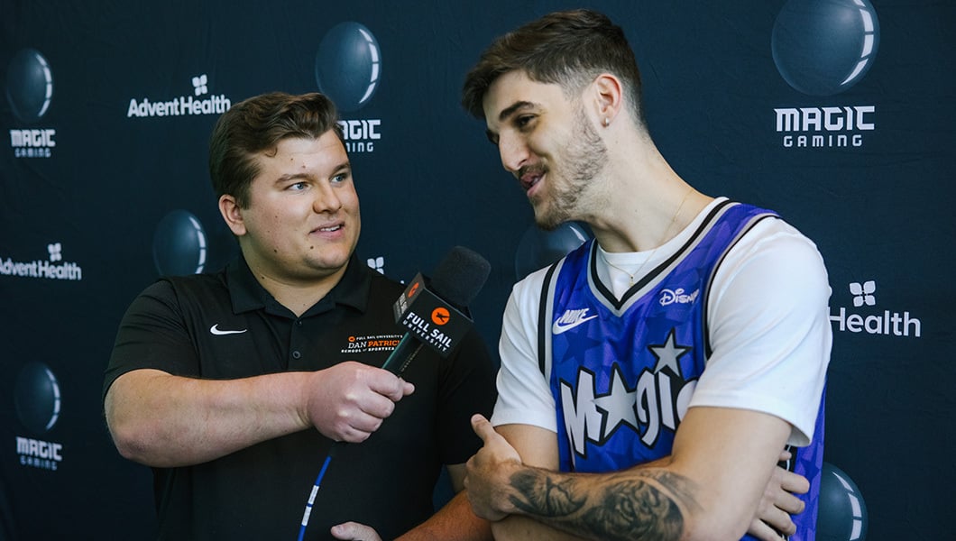 A person is holding a Full Sail branded microphone while interviewing a person in an Orlando Magic basketball jersey.