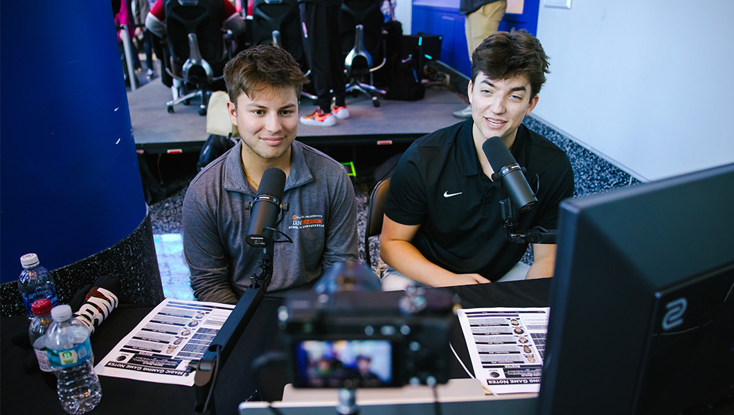 Two people are seated at a desk in front of microphones with a stage full of players in the background.