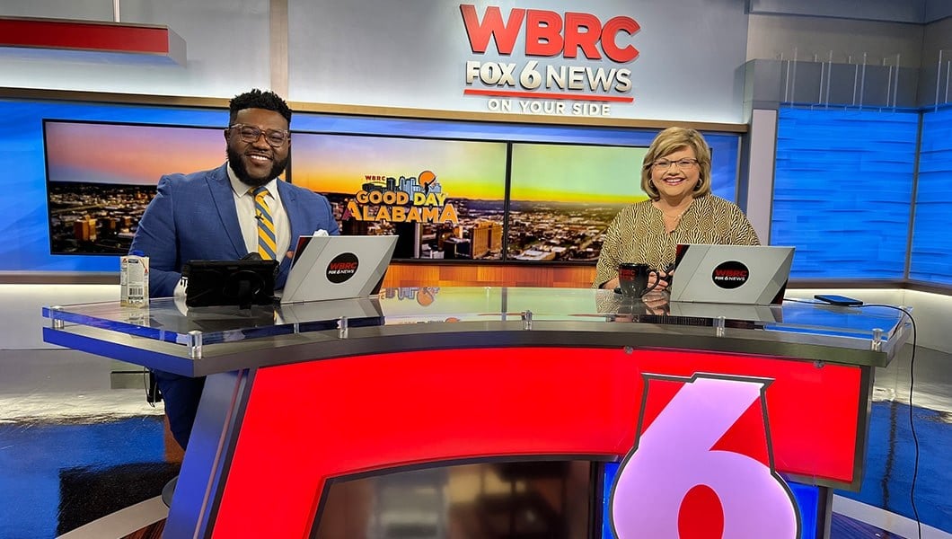 Toi Thornton sits at a news desk with his co-anchor. A screen behind them shows the Birmingham skyline and the Good Day Alabama logo.