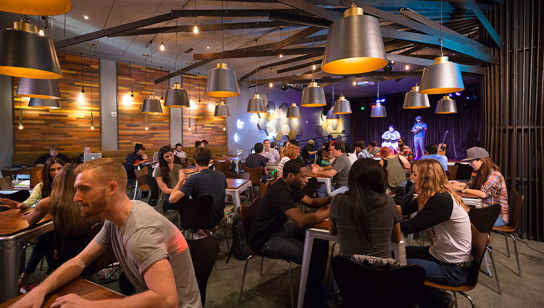 Students gather at tables for a lively event at the Treehouse, with a performance taking place on stage.
