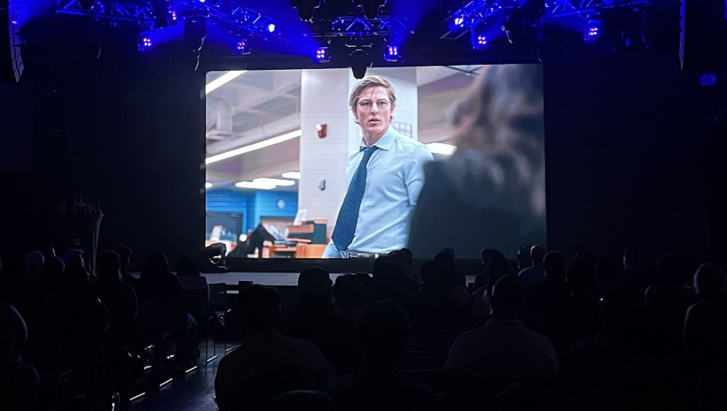 A large screen in a darkened room displays a character wearing a button-down shirt and tie.