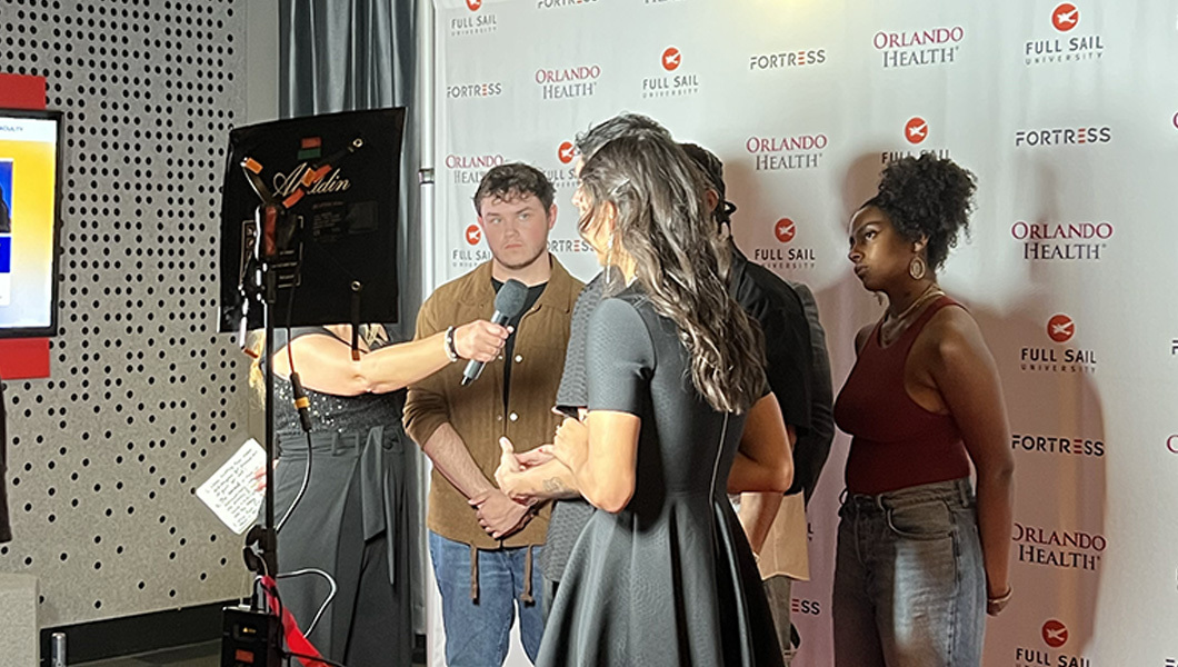 Several students stand on a red carpet in front of a background with the Fortress logo on it. An interviewer holds up a microphone to one student as a camera operator films them.