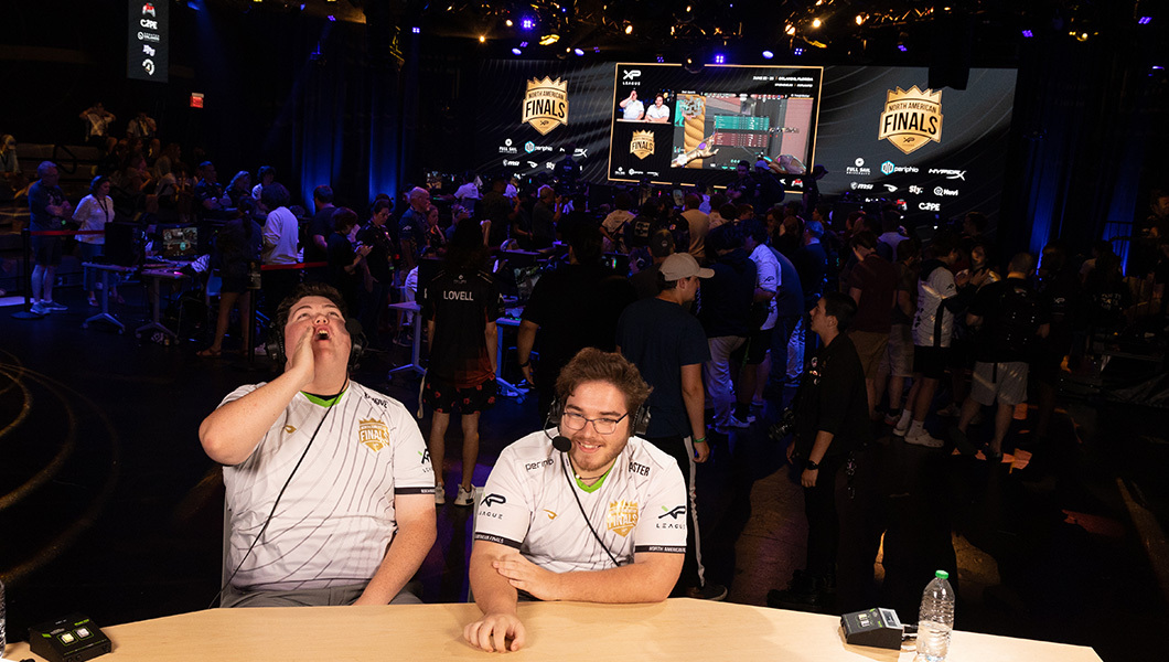 Two casters seated at a new desk while a large crowd observes the LED screen behind them.