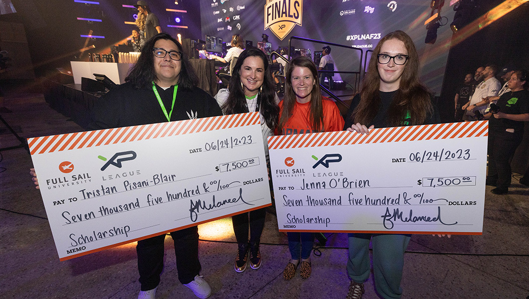 Four people standing to the side of a stage while holding large novelty checks and smiling.